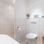 Badezimmer mit Toilette, Dusche und Badewanne. Weiße Fliesen und Holzregal.