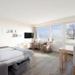 Zimmer mit Bett, TV, Esstisch und Balkon mit Blick auf Wasser.