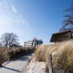 Weg durch Dünen mit Strohdachhaus und Hotels im Hintergrund.