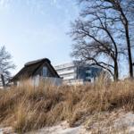 Dorfhaus mit Strohdach auf Dünen, moderne Gebäude im Hintergrund.
