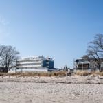 Strand mit Dünen, Bäumen und Ferienhäusern im Hintergrund unter blauem Himmel.