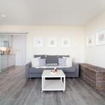 Wohnbereich mit grauem Sofa, weißen Kissen und Holzboden. Küche im Hintergrund.