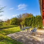 Terrasse mit Tisch und Stühlen neben roten Türen. Grünflächen und Bäume im Hintergrund.