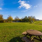 Holztisch und Bänke auf grünem Rasen. Spielplatz und Häuser im Hintergrund.