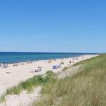 Strand mit Sanddünen und Menschen am Meer unter blauem Himmel.