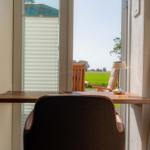 Schreibtisch mit Stuhl vor Fenster, Blick auf grünes Feld und Gartenmöbel
