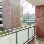 Terrasse mit Holzboden und Glasgeländer, Blick auf rote Backsteinhäuser und kahle Bäume.