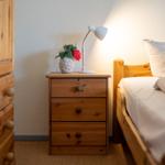 Schlafzimmer mit Holzbett, Nachttisch und Lampe. Auf dem Nachttisch steht eine Blumenvase.