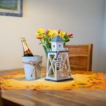 Holztisch mit Sonnenblumen-Tischdecke, Flasche Champagner und Strandturm-Dekoration.