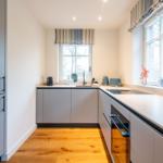 Küche mit grauen Schränken, Holzboden und Fenstern mit Rollläden.