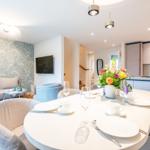 Esszimmer mit runder Tafel, Blumen und Sitzgelegenheiten. Küche und Wohnbereich im Hintergrund.