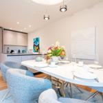 Essbereich mit Tisch und Stühlen, Küche im Hintergrund, Wandbilder und Blumen.