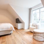 Schlafzimmer mit Bett, Fernseher und Sitzgelegenheit. Holzboden und großes Fenster.