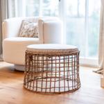 Rattan-Sitzhocker mit Kissen vor einem Sessel und Fenster