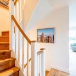 Holztreppenhaus mit Bild an der Wand und Blick in Wohnbereich mit Sitzgelegenheiten.