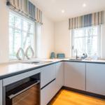 Küche mit hellen Schränken, Arbeitsplatte und zwei Fenstern mit Rollläden.