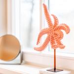 Ein orangefarbener Seestern auf einem Standfuß steht auf einem Fensterbrett neben einem runden Spiegel.