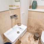 Badezimmer mit Waschbecken, Spiegel und Toilette. Wand und Boden sind mit hellen Fliesen ausgelegt.