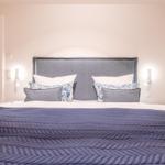 Doppelbett mit blauer Decke und Kissen, Wandlampen und Tür im Hintergrund.