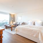 Ein Schlafzimmer mit Bett, Sofa, Tisch und Balkon. Holzfußboden und helle Wände.