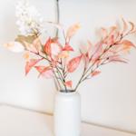 Ein weißer Vase mit Blättern und Blüten auf einer weißen Oberfläche.