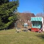 Grüner Rasen mit Strandkorb, Tisch und Stühlen. Hintergrund mit Bäumen und Häusern.