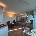 Schlafzimmer mit Bett, Sitzgelegenheit und Küchenbereich. Große Fenster und moderne Einrichtung.