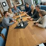 Gruppe sitzt um einen Holztisch mit Getränken. Blaue Sitzgelegenheiten und Teppich im Hintergrund.