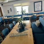 Großer Raum mit blauen Sitzgelegenheiten und Holztischen. Fenster und Wanddekoration sichtbar.