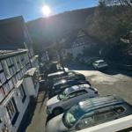 Parkplatz mit mehreren Autos vor traditionellen Häusern und bewaldeten Bergen unter Sonne.