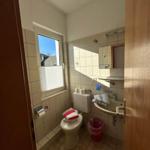 Badezimmer mit Toilette, Waschbecken und Fenster. Wandfliesen und roter Mülleimer.