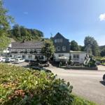 Traditionelles Haus mit Terrasse und Parkplatz vor grüner Landschaft.