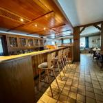 Bar mit Holzregalen und Sitzhöhenstühlen, Fliesenboden und Sicht auf Essbereich.