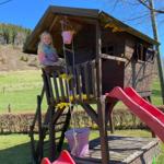 Ein Kind sitzt auf einer Holzspielburg mit roter Rutsche im Garten.