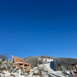 Holzstruktur auf schneebedecktem Hang mit Wald im Hintergrund unter blauem Himmel.