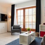 Wohnzimmer mit Sofa, Sessel, Fernseher und Balkonfenster.