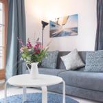 Ein grauer Sofa mit Kissen, weißer Tisch und Blumen vor Fenster.