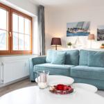 Ein Wohnzimmer mit blauem Sofa, weißen Tischen und Fenstern mit Vorhängen.