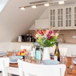 Essbereich mit Tisch, Stühlen und Blumenarrangement. Küche mit Schränken und Arbeitsfläche.