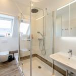 Badezimmer mit Dusche, Waschbecken und WC. Fenster mit Blick nach außen.