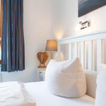 Schlafzimmer mit weißem Bett, Nachttisch, Lampe und Fenster mit Vorhängen.