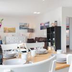 Essbereich mit Tisch und Stühlen, Blumen und Flasche. Im Hintergrund Wohnzimmer und Schlafzimmer.