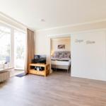 Wohnzimmer mit Schlafzimmer dahinter. Große Fenster, Holzboden und TV.