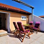 Terrasse mit Holztisch und vier Stühlen mit lila Sitzkissen. Tür führt ins Haus. Hintergrund: Haus mit rotem Dach.