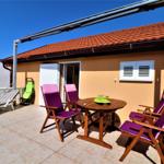 Terrasse mit Tisch, vier Stühlen und Liegestuhl unter Sonnenschutz.