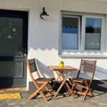 Vor dem Eingang steht ein Tisch mit zwei Stühlen. Eine Tür und ein Fenster sind sichtbar.