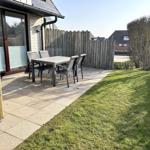 Terrasse mit Tisch und Stühlen, Grasfläche und Holzzaun. Wickermöbel links.