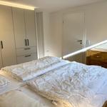 Schlafzimmer mit Bett, Kleiderschrank und Nachttisch. Sonnenlicht fällt durch Fenster.