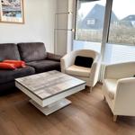 Wohnzimmer mit Sofa, zwei Sesseln und Couchtisch. Große Fenster mit Blick auf Außengebiet.