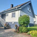 Haus mit Eingangstreppe, Blumenbeete und gepflastertem Vorplatz unter blauem Himmel.